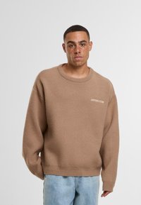 Béžový oversized svetr vyrobený z měkkého materiálu s kulatým výstřihem a žebrovanými manžetami; na hrudi se nachází malé bílé logo.