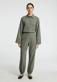 Olijfgroene tweedelige outfit bestaande uit een knoopshirt met een zak en wijde broek met elastische boorden. Glad stoftextuur.