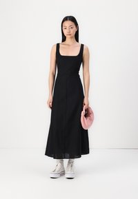 Rochie neagră fără mâneci, cu decolteu pătrat, tiv fluid și textură netedă. Asortată cu o poșetă roz și adidași albi cu glezna înaltă.