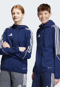 Marineblaue Jacken mit weißen Akzenten und drei Streifen. Verfügt über einen Reißverschluss, Kapuze und atmungsaktives Material. Adidas-Logo sichtbar.