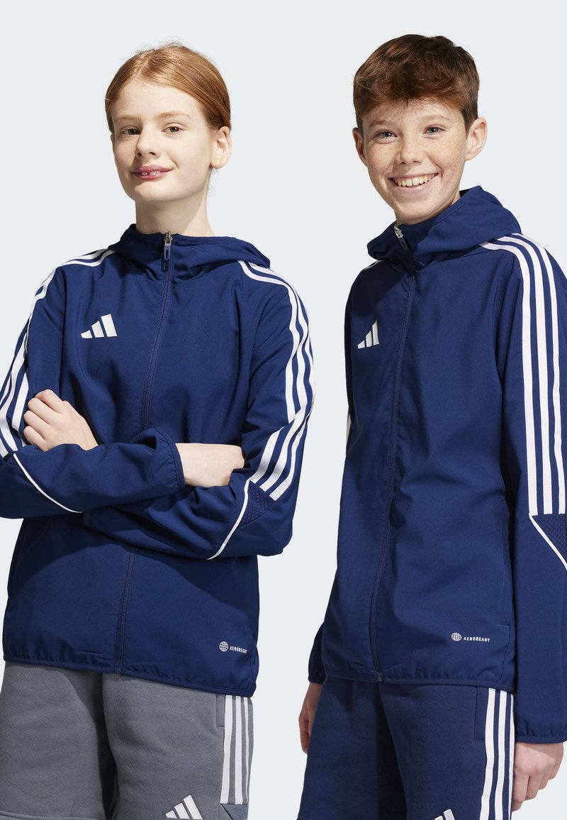 Marineblaue Jacken mit weißen Akzenten und drei Streifen. Verfügt über einen Reißverschluss, Kapuze und atmungsaktives Material. Adidas-Logo sichtbar.