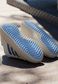 Bijele tenisice s plavim cik-cak potplatom, tri plave pruge sa strane, teksturirani potplat i zaobljen oblik. Nema vidljivog brendiranja na stranama.