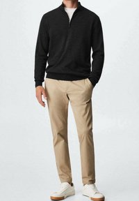 Czarny sweter rozpinany na zamek, beżowe chinosy i białe trampki. Sweter ma wysoki kołnierz i ściągacze, a chinosy są zwężane.