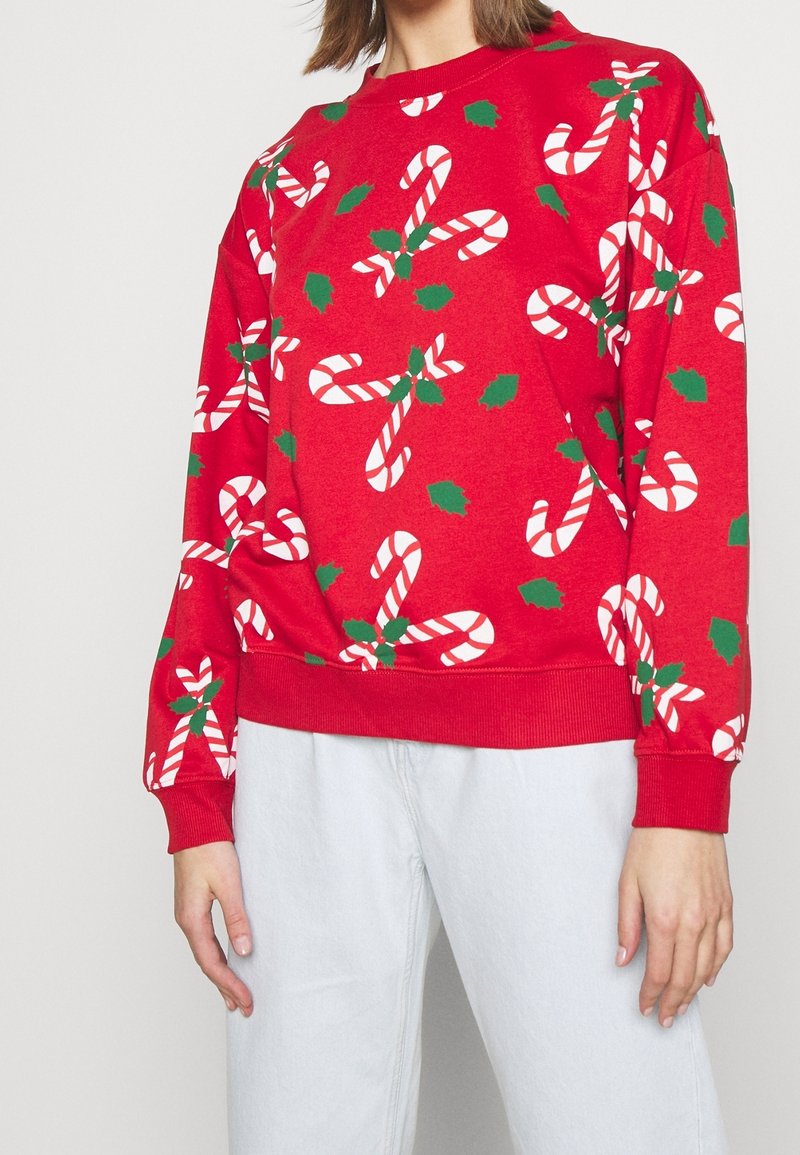 Pull rouge à manches longues avec des motifs de cannes en bonbon blanches et rouges et des feuilles de houx vertes, porté avec un pantalon bleu clair.