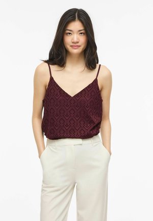 Kobieta o długich ciemnych włosach, ubrana w bordową, wzorzystą bluzkę na cienkich ramiączkach i białe spodnie z wysokim stanem, ręce w kieszeniach, neutralne tło.