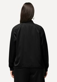 Giacca in tessuto nero e liscio con un fitting rilassato e polsini a coste. Presenta un design della schiena dritta con hardware minimale e senza loghi visibili.