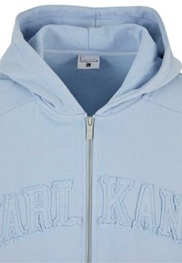 Lyseblå zip-up hættetrøje med broderet "KARL KANI" tekst, hætte, ribbet tekstur og sølv lynlås. Blødt stof og afslappet design.