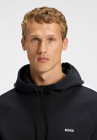 Black zip-up hoodie featuring a drawstring hood, smooth fabric, white accent on shoulders, and "BOSS" logo on the chest.