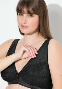 Czarny koronkowy biustonosz braletka z dekoltem w kształcie litery V. Posiada regulowane ramiączka, dekoracyjny guzik na środku oraz teksturowany, wzorzysty materiał.