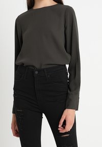 Long-sleeve, dark green blouse with a crew neck, paired with black distressed jeans featuring ripped details and a high-waisted fit.