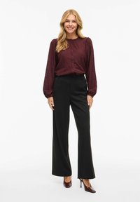 Smilende kvinde med langt lyst hår iført en bordeaux tekstureret bluse, sorte bukser med brede ben og bordeaux højhælede sko.