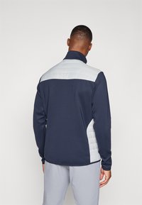 Jūras spēku zips jaka ar strukturētu gaiši pelēku mīkstu aizmugures paneli, mīkstu audumu un augstu apkakli, kas paredzēta komfortam un kustību brīvībai.