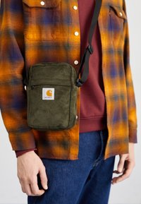 Henkilö, jolla on päällään oranssi ja sininen ruudullinen paita marooninvärisen collegepaidan päällä, vihreä corduroy Carhartt -ristilaukku ja siniset farkut.