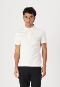 Polo shirt en coton blanc avec un col classique, quatre boutons, des manches courtes et un petit logo sur la poitrine gauche. Porté avec un pantalon noir.