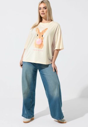 Donna che indossa una maglietta crema oversize con un coniglio che soffia una gomma da masticare rosa e jeans a gamba larga blu, in piedi davanti a uno sfondo semplice.
