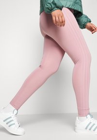 Ljusrosa leggings med en slät yta, som har två vertikala ränder på sidan. Kombination med vita sneakers och en grön jacka.