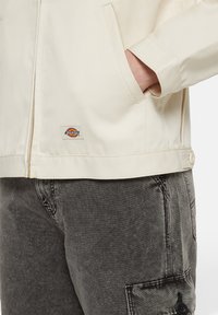 Beige lightweight jacket with zip closure and side pockets, featuring a small Dickies logo patch at the hem. Worn over dark gray cargo pants.