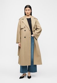 Γυναίκα που φοράει μπεζ καμπαρντίνα, μαύρο τοπ, τζιν μπλε με φαρδιά πόδια και μαύρα παπούτσια με τακούνι, στέκεται με το ένα χέρι στην τσέπη.