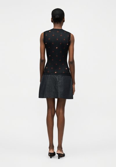 Γυναίκα με τα μαλλιά σε κότσο φοράει αμάνικη μαύρη μπλούζα με κόκκινο σχέδιο κερασιού, σκούρα φούστα και μαύρες γόβες, στέκεται απέναντι σε λευκό φόντο.