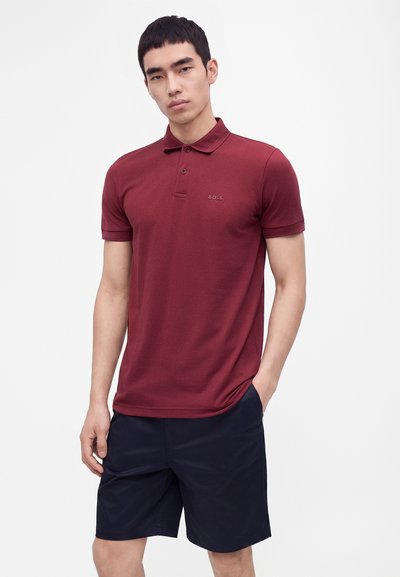 Homme portant un polo bordeaux à manches courtes et un short noir, debout sur un fond blanc uni.