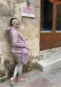 Felpa rosa chiaro con tasca laterale, leggings pastello abbinati e una borsa a tracolla frangiata. Lo sfondo presenta muri di pietra e porte di legno.