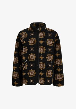 Fekete mintás polárdzseki barna részletekkel, narancssárga kapucnis póló fölé húzva. Laza szürke nadrág és bézs cipő teszi teljessé az öltözéket.