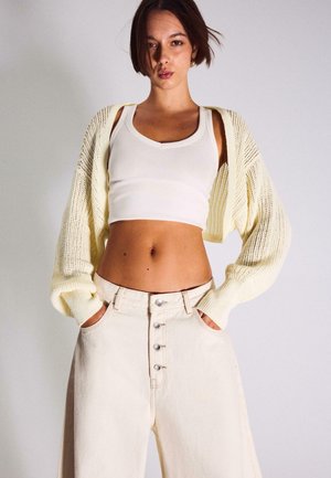 Femme portant un débardeur court blanc, un cardigan en maille crème, et un pantalon large crème à taille haute avec boutons, les mains dans les poches.