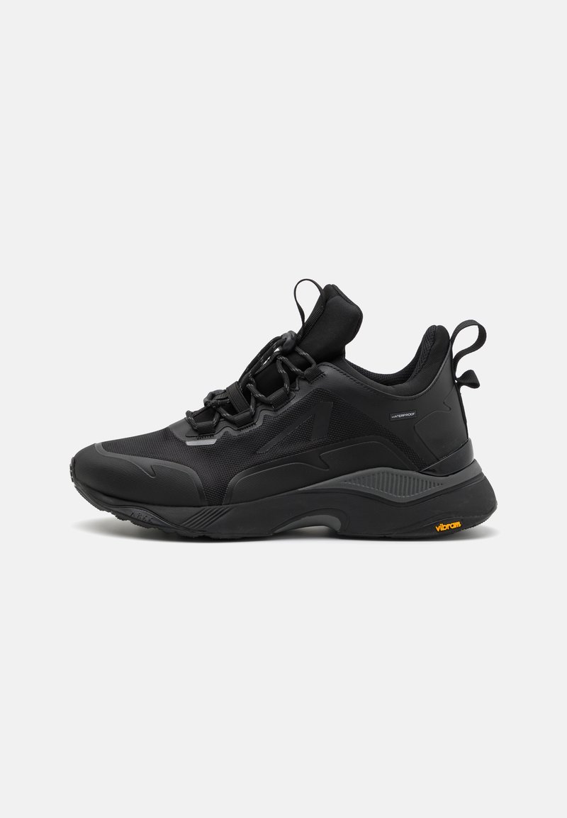 Chaussure de sport noire avec une tige en mesh respirant, une semelle épaisse en caoutchouc, des détails texturés et un logo Vibram bien visible sur la semelle extérieure. Conception imperméable.