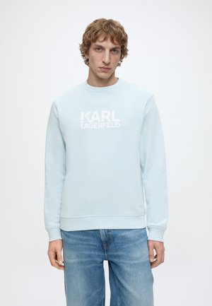 Jonge man met golvend haar, dragende een lichtblauwe sweatshirt met "KARL LAGERFELD" logo, gecombineerd met blauwe spijkerbroek, staand tegen een effen achtergrond.