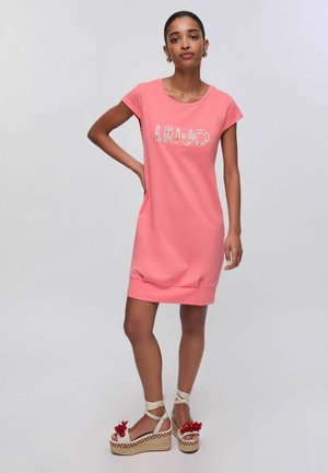 Giovane donna con capelli scuri che indossa un abito rosa corallo con la scritta "LIU JO" e sandali beige con zeppa e decorazioni floreali rosse.