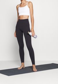 Czarne legginsy z wysokim stanem i biały skrócony top. Osoba trzyma wzorzysty bidon, stojąc boso na szarym macie do jogi.