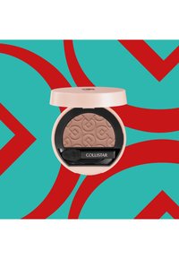 Poudre compacte dans un boîtier rond beige. Présente une surface texturée et embossée de couleur brun clair. Comprend une éponge applicatrice noire.