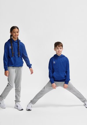Blå hættejakke med frontlomme og logo, parret med grå sweatpants. Begge børn har hvide sportssko på. Ensfarvet baggrund.