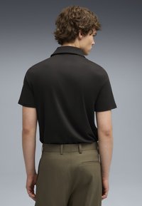 Polo shirt negru cu mânecă scurtă și guler clasic, purtat cu pantaloni de tip olive. Textură netedă și croială ajustată.