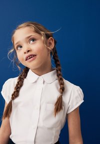 Bela kratka srajca s kratkimi rokavi, za zapenjanje s srajčnimi gumbi, z zaobljenim ovratnikom in žepom, kombinirana s temno krilom, na modrem ozadju.