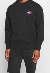 Czarna bluza z żebrowanym, okrągłym dekoltem i elastycznymi ściągaczami, z małym logo "TOMMY JEANS" w kolorach czerwonym, białym i niebieskim na piersi.