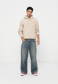 Beige Fleece-Hoodie mit Kängurutasche, kombiniert mit weitgeschnittenen blauen Jeans. Die Schuhe sind rot-weiße Sneaker.