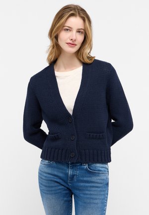 Cardigan en maille bleu marine avec un col en V, des poignets côtelés, deux poches avant et une fermeture à boutons, associé à une chemise légère et un jean.