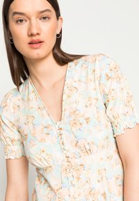 Vestido floral com tons de azul pastel e pêssego, mangas curtas com volume e decote em V. Apresenta botões decorativos na parte da frente e um tecido suave.