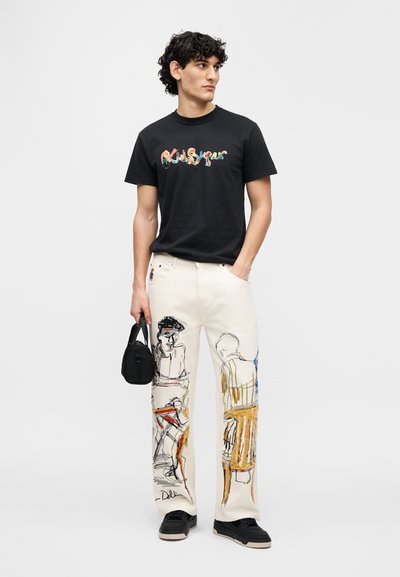 Μαύρο βαμβακερό μπλουζάκι με πολύχρωμο γραφικό κείμενο· μπεζ παντελόνια με καλλιτεχνικά σκίτσα. Μαύρη τσάντα και μαύρα παπούτσια συμπληρώνουν το σύνολο.