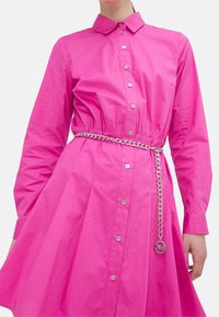 Abito camicia rosa in cotone, con chiusura a bottoni sul davanti, maniche lunghe e una cintura in catena argentata con un emblema circolare.