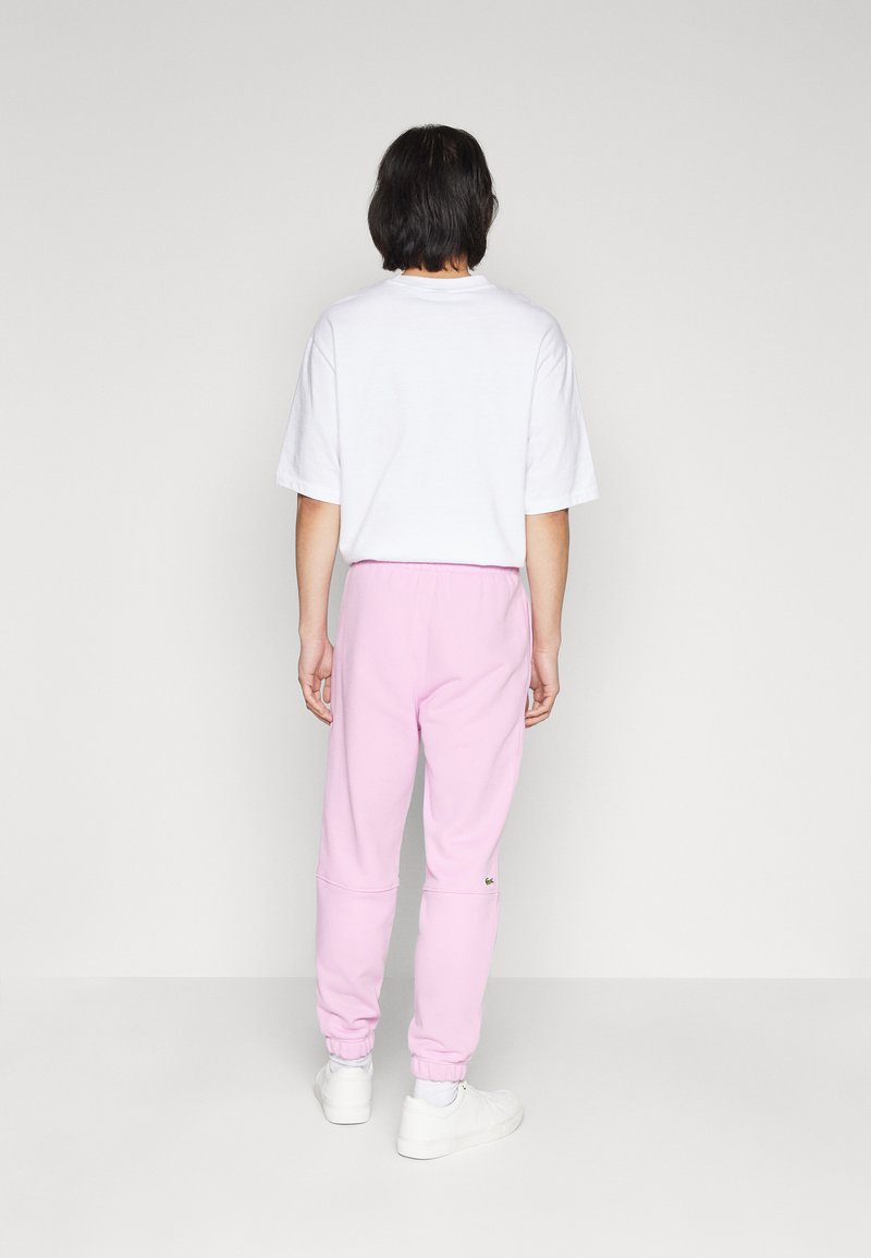 Camiseta blanca de manga corta y pantalones de chándal rosa con puños elásticos. Los pantalones de chándal cuentan con un pequeño detalle de logo cerca del tobillo izquierdo.