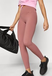 Ροζ παντελόνι κολάν σε απόχρωση βατόμουρου με ψηλή μέση, λεία υφή και ραφές flatlock. Συνδυάζεται με μια μαύρη τσάντα duffel και μαύρα αθλητικά παπούτσια.