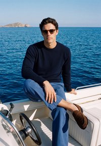 Jeune homme portant des lunettes de soleil, un pull bleu marine et un pantalon bleu, assis avec une jambe croisée sur un bateau blanc avec l'océan et une île en arrière-plan.