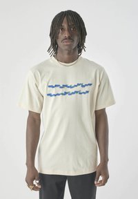 Man with dreadlocks wearing a cream T-shirt with blue and green cartoon caterpillars printed in two horizontal lines across the chest.