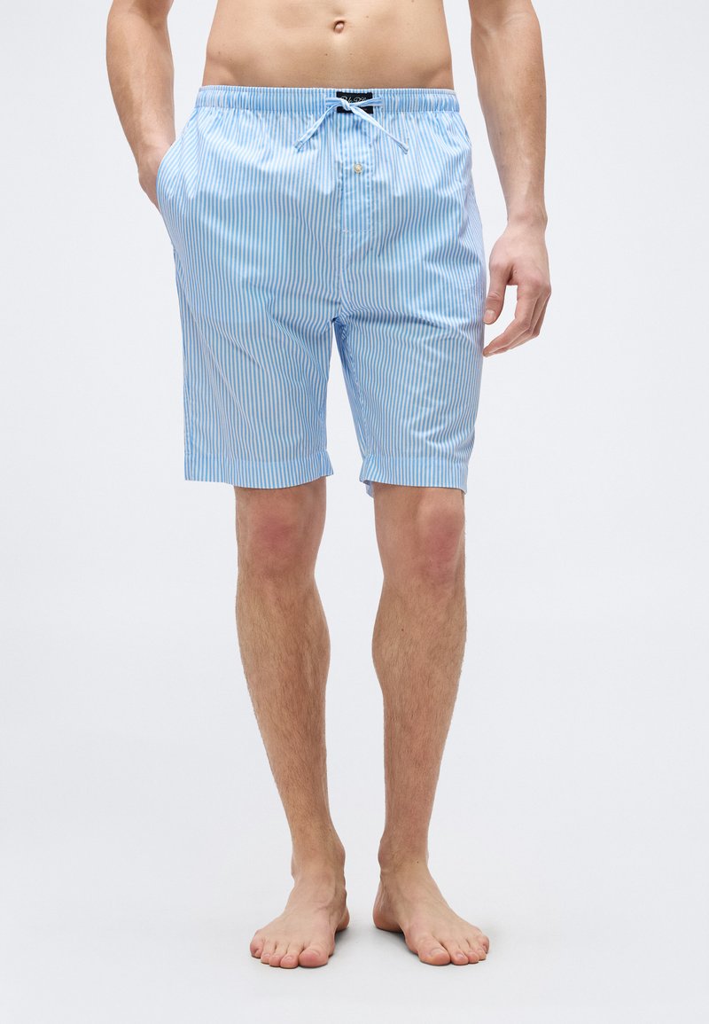 Άνδρας με ανοιχτό μπλε και λευκή ριγέ σόρτς με κορδόνι, όρθιος ξυπόλυτος με τα χέρια στις τσέπες σε απλό φόντο.