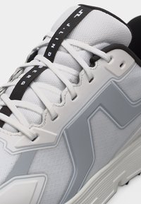 Κοντινό πλάνο ενός λευκού και γκρι αθλητικού παπουτσιού με ύφασμα από δίχτυ, λεπτομερές ράψιμο, λευκά κορδόνια και μαύρη θηλιά με την ένδειξη "THUNDER".