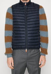 Marineblå puffer vest med vandret syninger, høj krave og en frontlynlås med en orange trækstrop, båret over en stribet sweater.