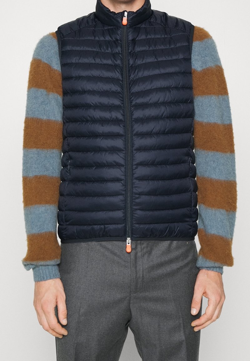 Marineblå puffer vest med vandret syninger, høj krave og en frontlynlås med en orange trækstrop, båret over en stribet sweater.