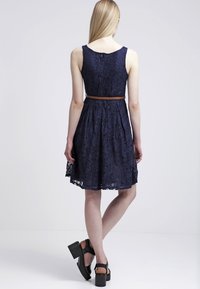 Navy blauwe kanten jurk met een aansluitende top en een uitlopende rok, voorzien van een bruine riem in de taille. Gladde textuur, knielange snit.